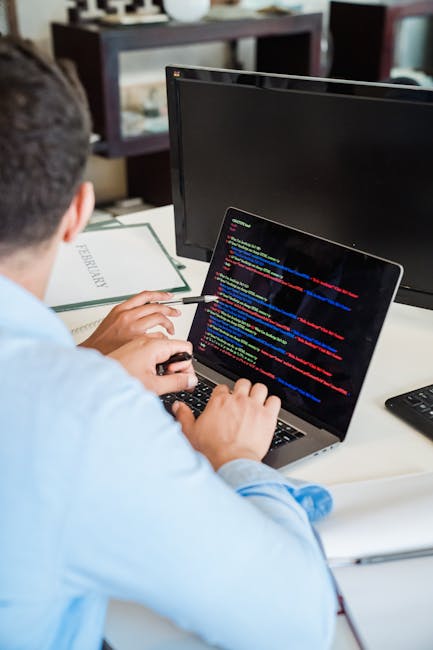 A man with glasses and a beard is working at a wooden desk with two large computer monitors displaying lines of code and programming software. The left monitor shows a dark-themed interface with folde