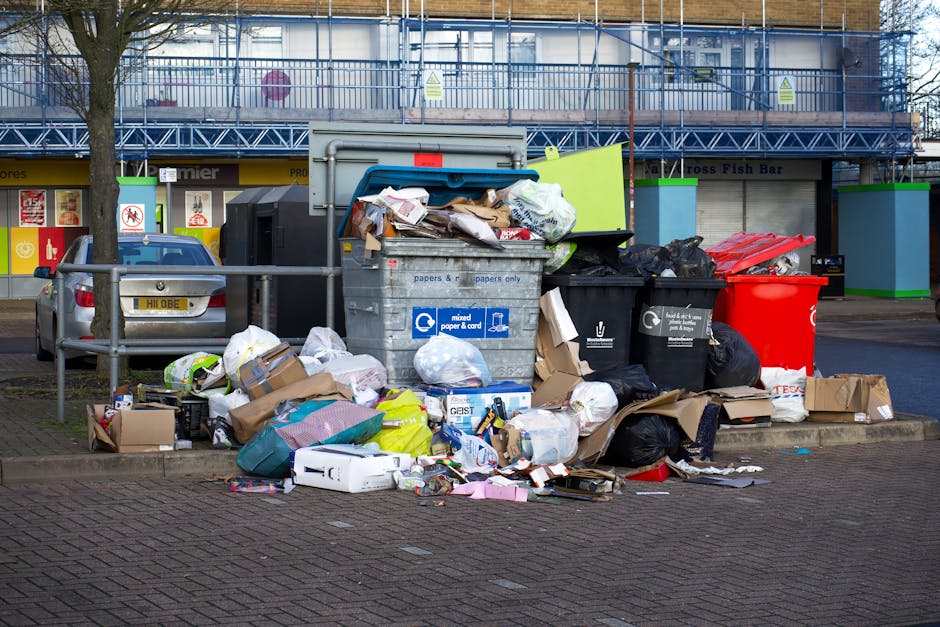 A sanitation worker, wearing a yellow high-visibility vest with red accents, is operating a large red waste collection vehicle on a street. The back of the vehicle has an open compactor section with v